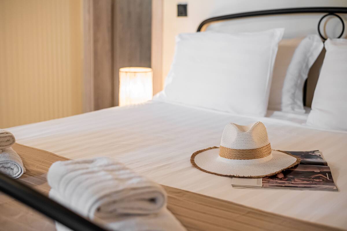 Un lit bien fait, avec des draps blancs, une serviette enroulée, un chapeau de paille et un magazine posés sur le lit.