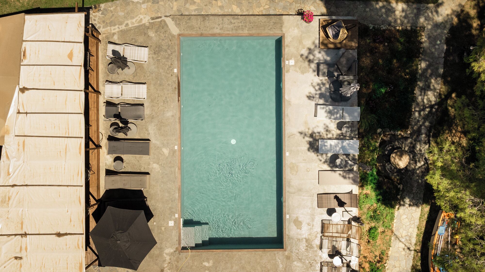 An aerial view of a rectangular swimming pool surrounded by lounge chairs and umbrellas, with a stone wall on one side and a pathway leading to a building.