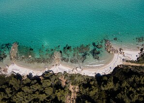 An aerial view of a beach with clear turquoise waters, sandy shore, and dense forested area. - Pefkon Suites (Kassandra)