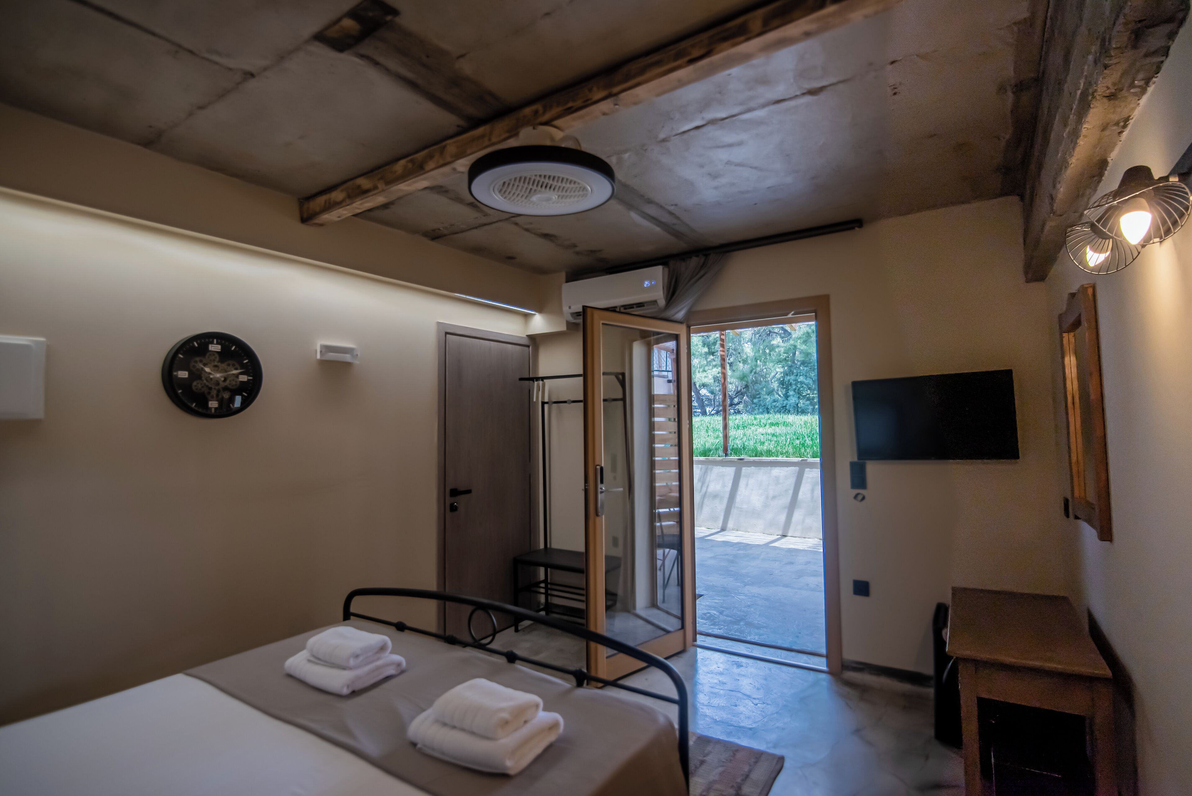 A bedroom with a bed, a clock, an air conditioning unit, a television, and a sliding door leading to an outdoor area.