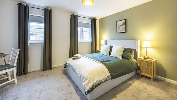 A bedroom with a bed, bedside tables, a chair, and a window with curtains. - The Lake Hub (Dagenham)