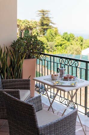 Een balkon met rieten meubels, een tafel met koffiepot, een schaal koekjes en uitzicht op zee.