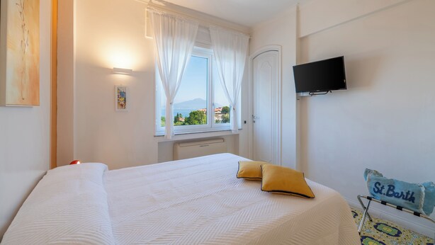 A hotel room with a bed, a TV mounted on the wall, a window with curtains, and a view of the sea. - Amazing apt with breathtaking seaview&Vesuvius in Sorrento center near the beach (Sorrento)