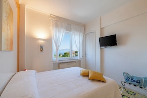 Ein Hotelzimmer mit Bett, einem an der Wand befestigten Fernseher, einem Fenster mit Vorhängen und Meerblick.