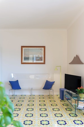 Een kamer met een transparante plastic stoel, een lijst met een afbeelding aan de muur, een televisie en een vloer met een patroon.