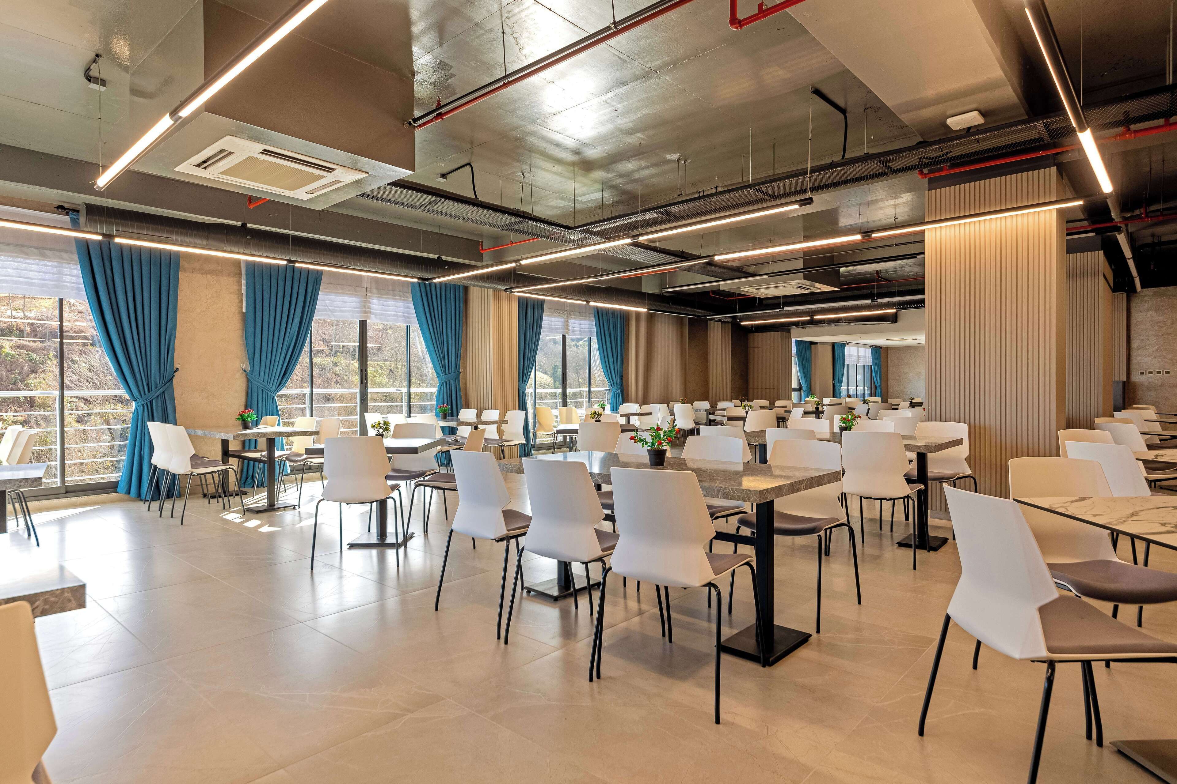 A spacious dining area with multiple tables and chairs, large windows with blue curtains, and a modern ceiling with exposed pipes and lighting.