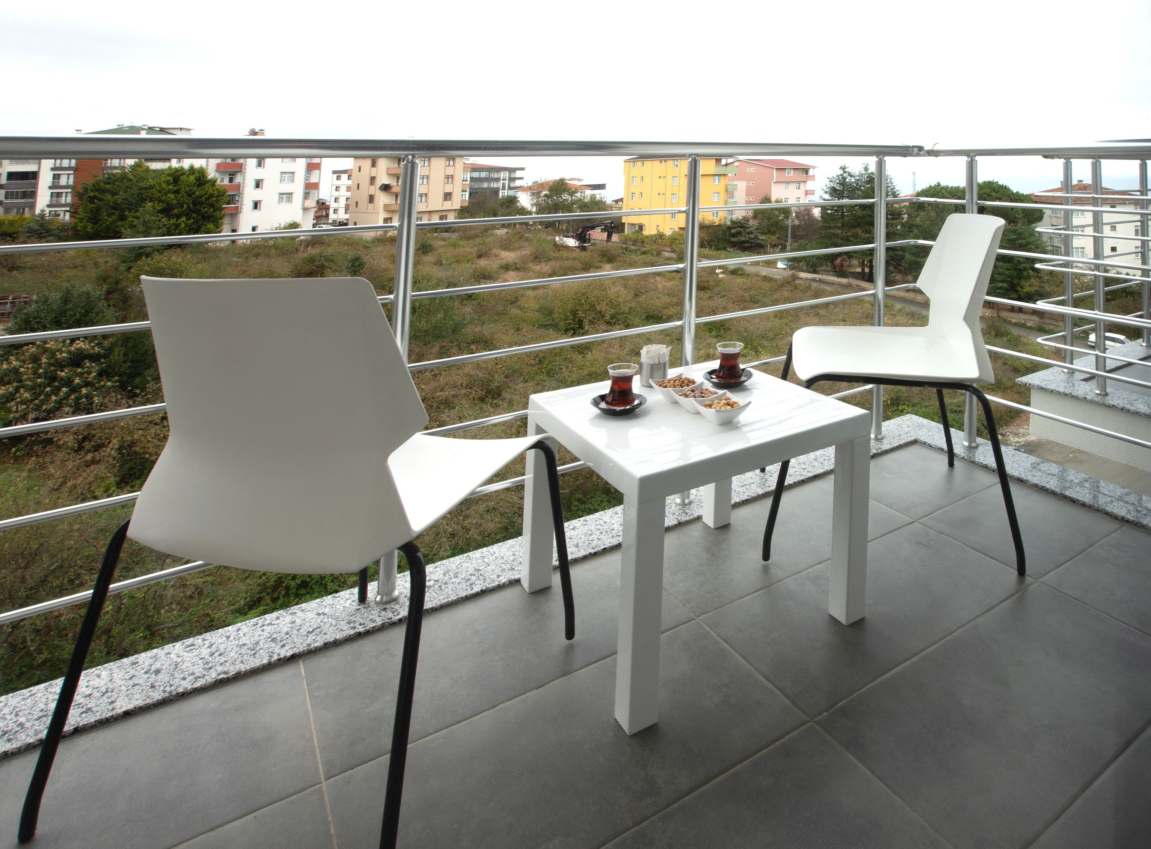 A balcony with two white chairs and a table, a small tray with snacks, and a view of buildings and greenery.