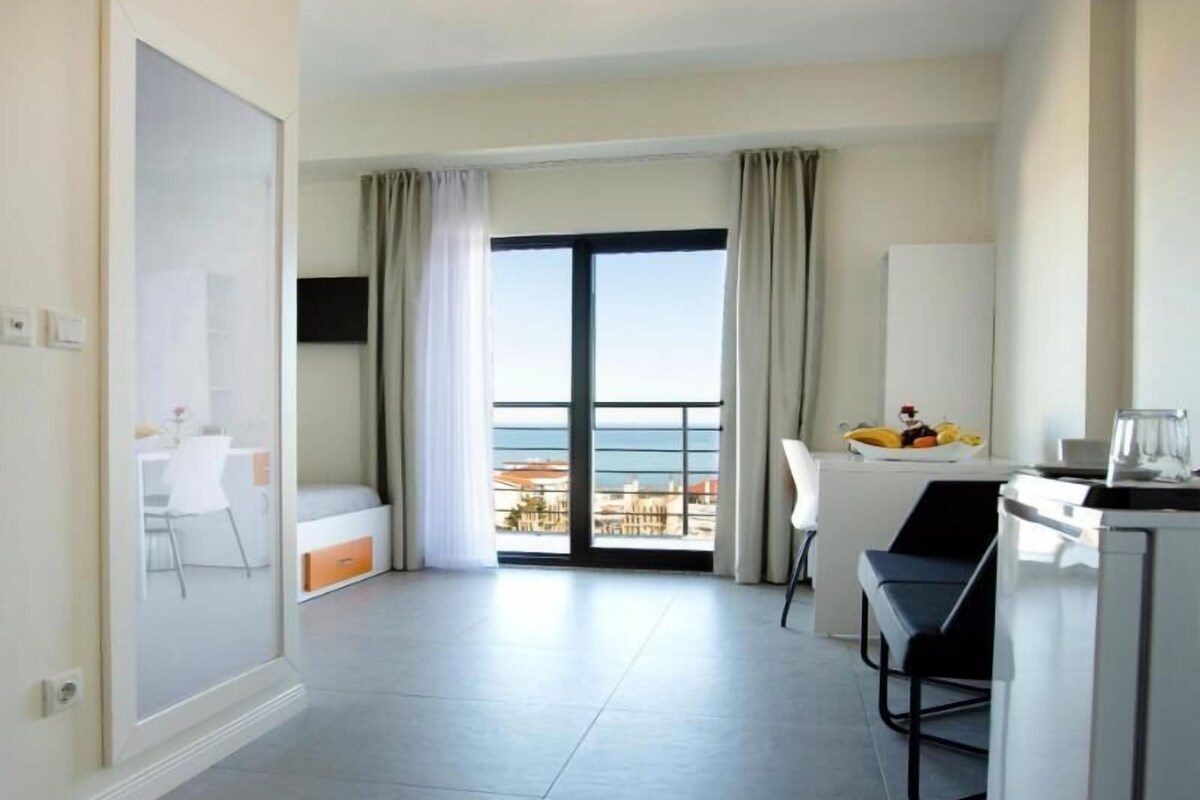 A modern hotel room with a balcony, a dining area, and a kitchenette.
