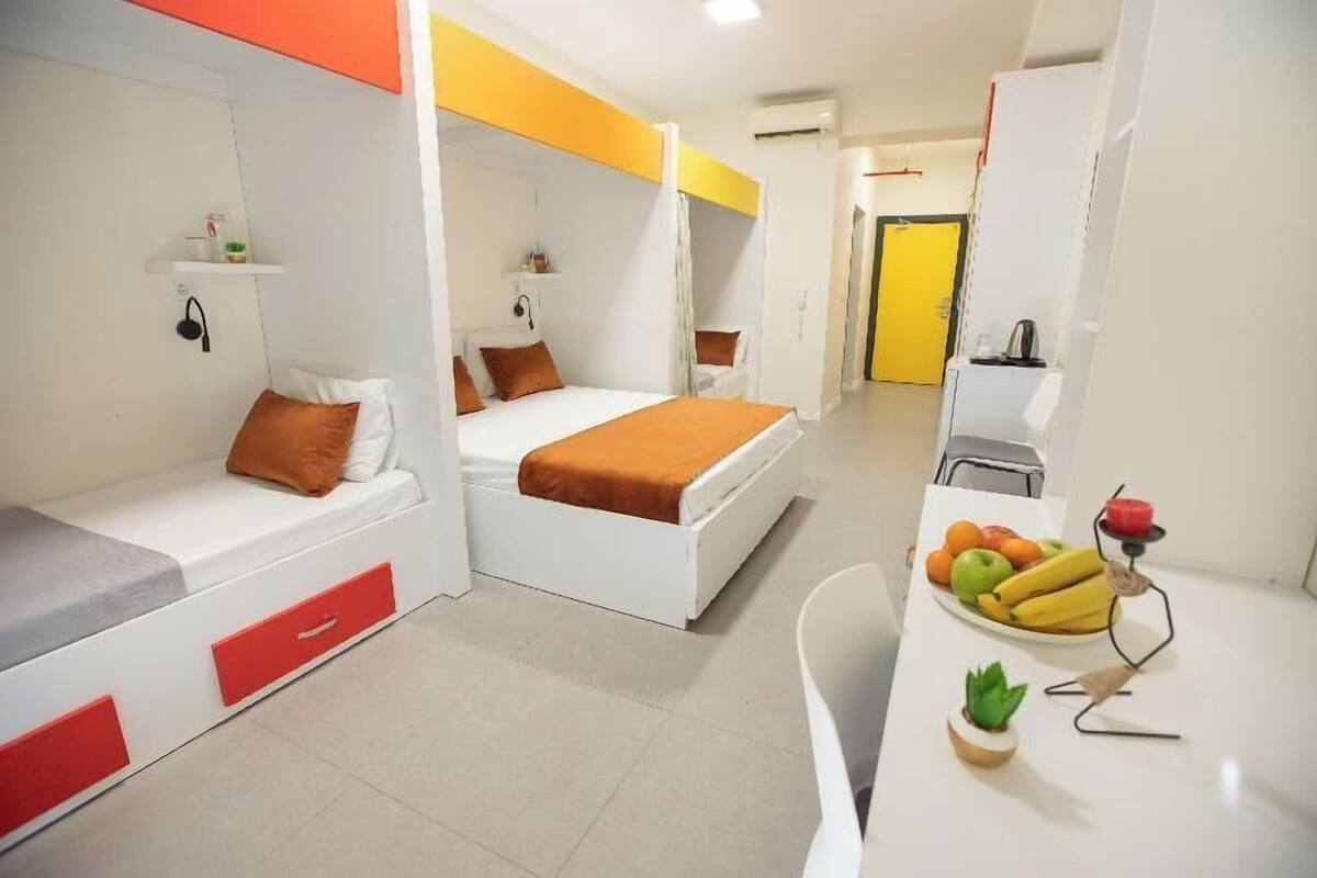 Een compacte hotelkamer met een bed, een klein bureau met een schaal fruit en een kitchenette.