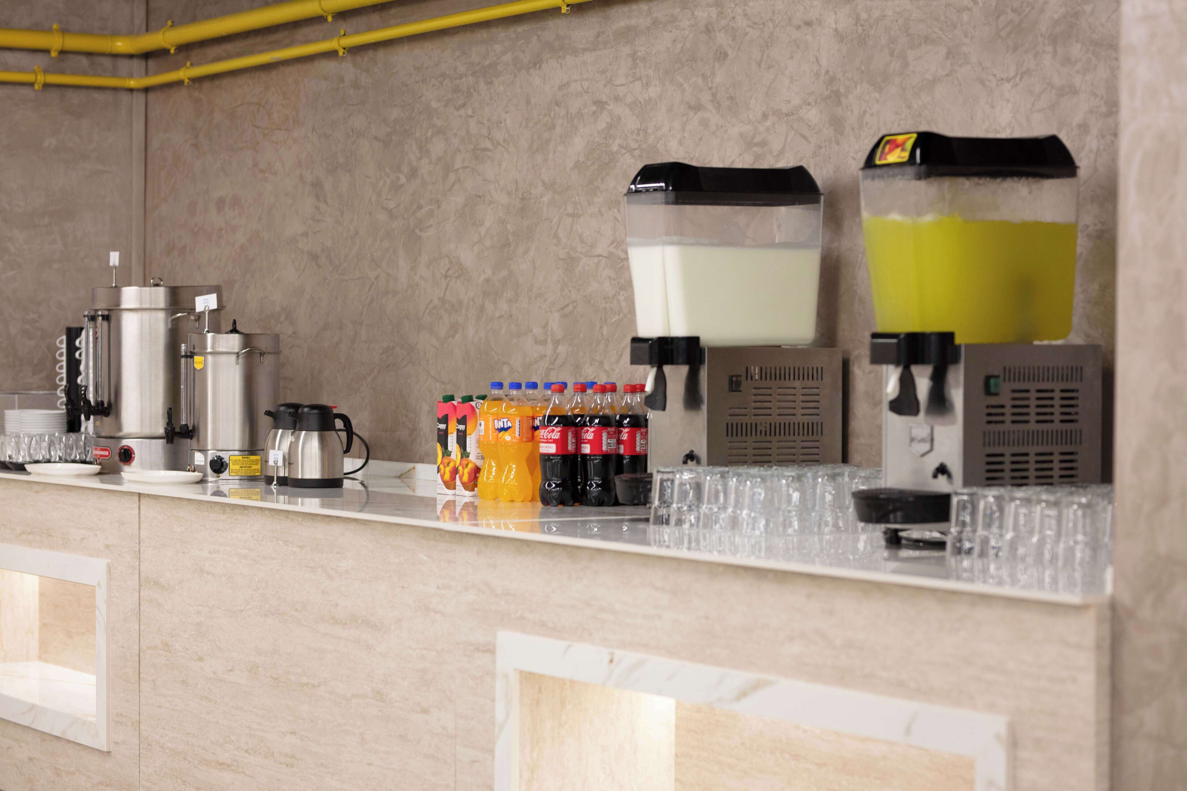A bar area with a coffee machine, a milk dispenser, and a soda dispenser. There are also several bottles of soda and a stack of glasses on the counter.