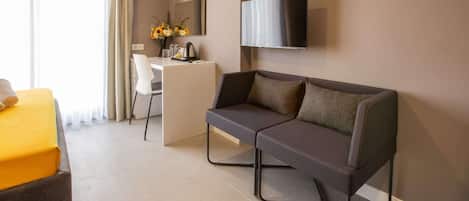 A modern hotel room with a sofa, a desk, a chair, a TV mounted on the wall, and a window with curtains.
