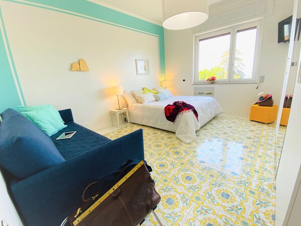 Ein Zimmer mit einem Bett, einem blauen Sofa, einem kleinen Tisch und einem Fenster mit Blick auf Grünflächen.
