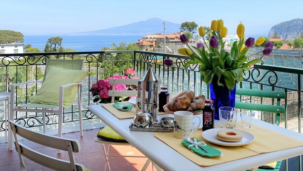 Uma varanda com uma mesa posta para o café da manhã, incluindo croissants, uma garrafa de suco e um vaso de flores.