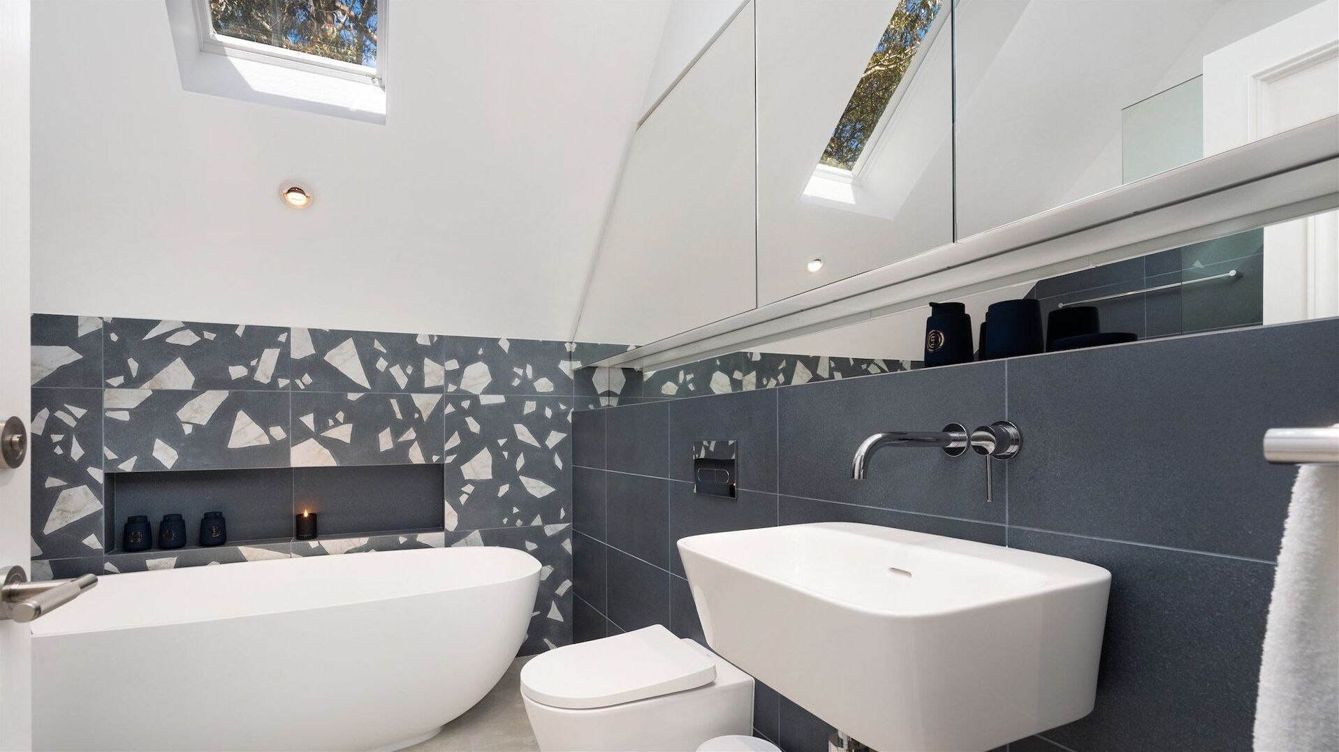 A modern bathroom with a freestanding bathtub, a pedestal sink, and a toilet. There is a skylight above the bathtub and a large mirror above the sink.