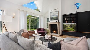 Een moderne woonkamer met een open haard, een televisie en een groot raam met uitzicht op een tuin.