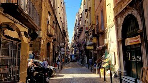 A cobblestone street lined with multi-story buildings, some with balconies and outdoor seating. - Tribunali Platamon (Naples)