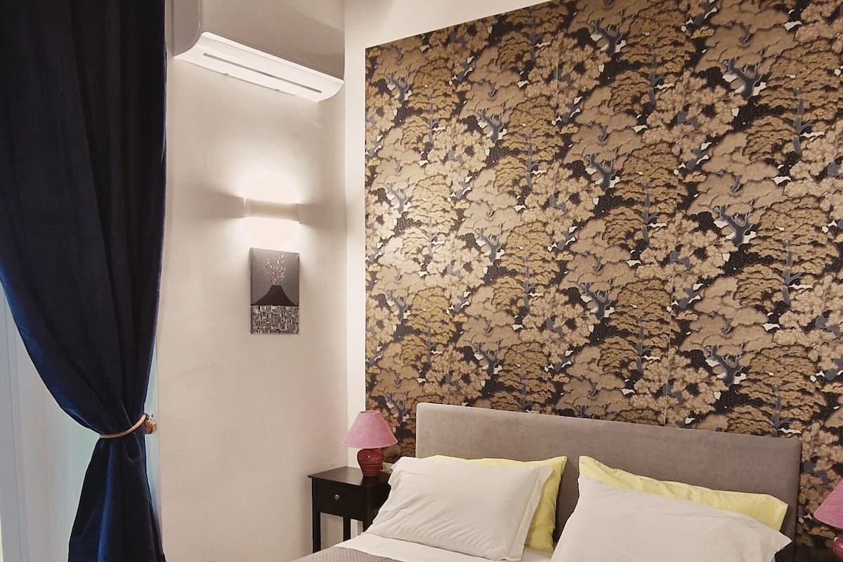 A bedroom with a bed, a nightstand, a lamp, a wall-mounted artwork, and a window with curtains.
