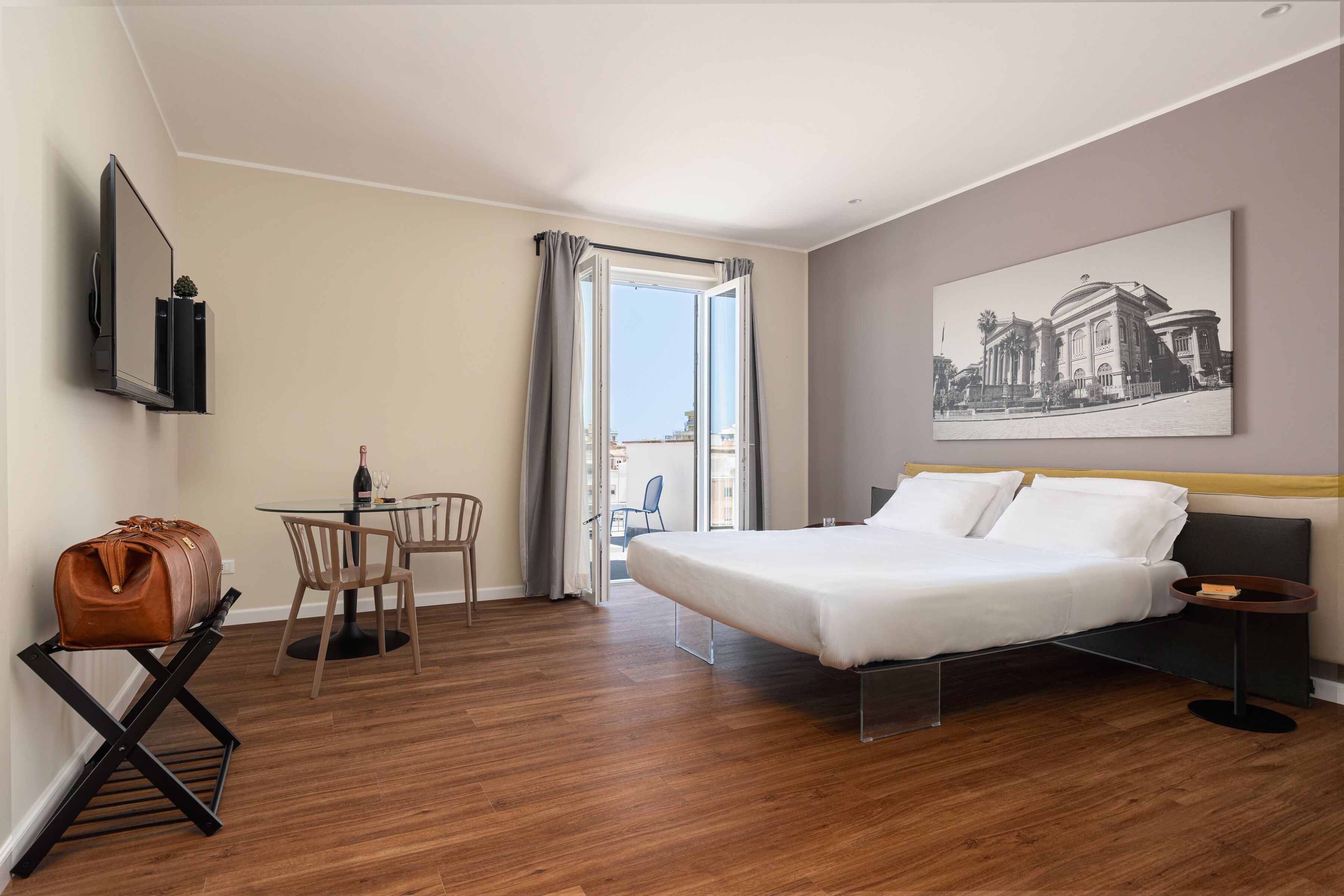 A hotel room with a bed, a desk, a chair, a wine bottle, and a view of a building through a window.