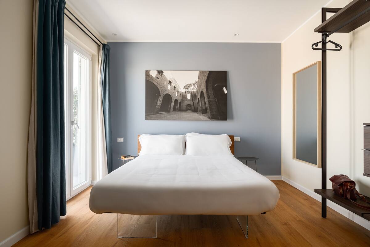 Un dormitorio con una cama grande, una ventana con cortinas, un cuadro en la pared y una mesita de noche.