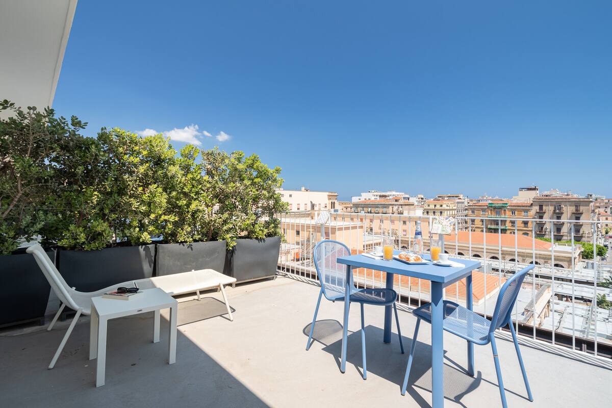 Un balcón con una mesa blanca y sillas azules, con vistas a un paisaje urbano.