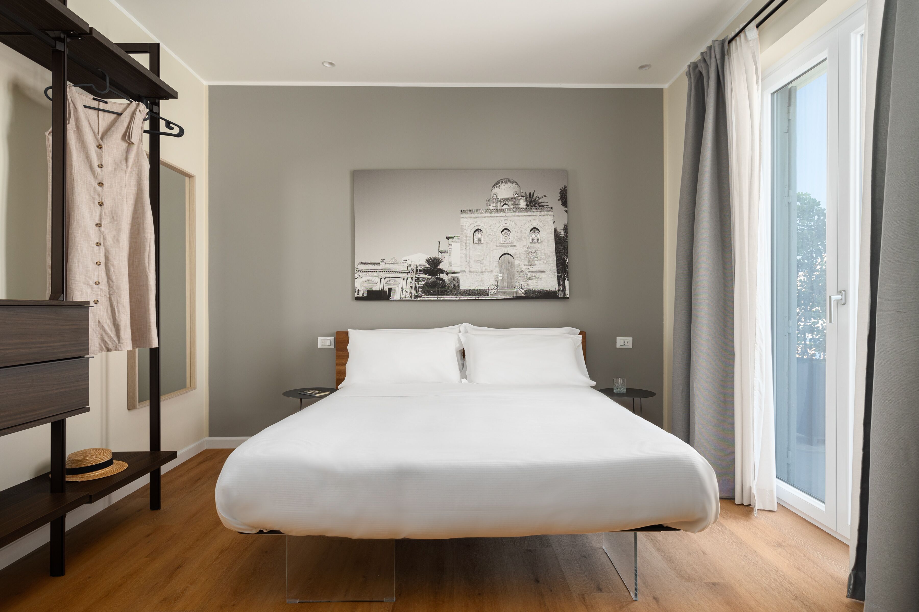 Ein modernes Hotelzimmer mit einem großen Bett, Holzboden, einem Wandbild, einem Nachttisch und Blick ins Freie durch ein Fenster.