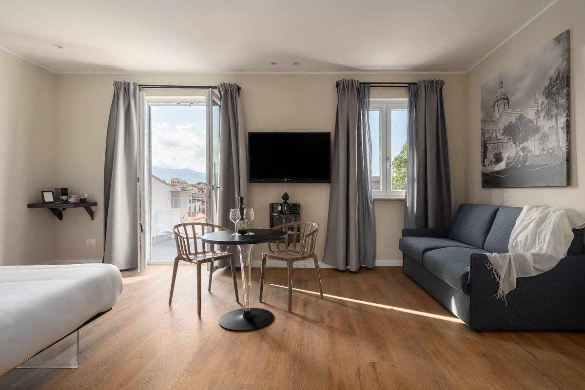 Una sala de estar moderna con un sofá, una mesa de comedor con sillas, un televisor en la pared y un balcón con cortinas.