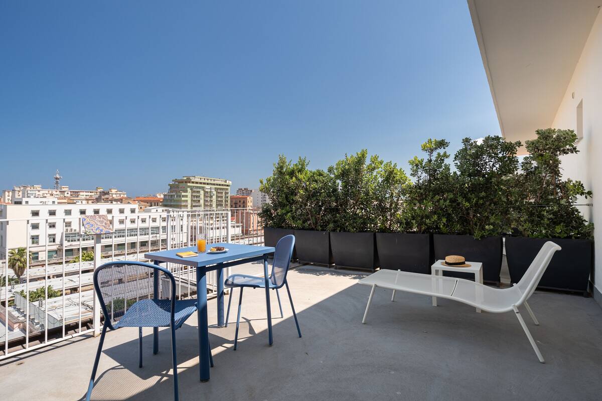 Un balcón con mesa y sillas, una silla de descanso blanca y vistas a edificios y árboles.
