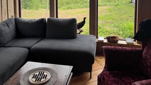 Una habitación acogedora con un sofá seccional gris oscuro, una mesa de centro de madera con un cuenco decorativo, y vistas a un paisaje verde exuberante a través de amplias ventanas.