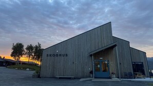 Un edificio de madera con el nombre "SKOGHUS" en su fachada, que presenta una puerta grande y un pequeño porche con asientos.