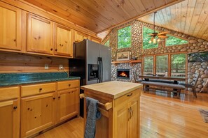 A wooden cabin kitchen with stone walls, a stone fireplace, and a large dining table.
