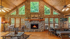 A cozy cabin interior with a stone fireplace, wooden furniture, and large windows offering views of the forest. - Sunset in the Mountains – Blue Ridge, GA. (Blue Ridge)