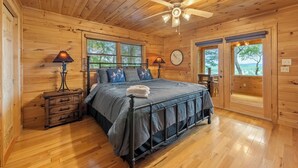 A wooden cabin bedroom with a bed, two bedside lamps, a nightstand, a ceiling fan, and a view through the door. - Sunset in the Mountains – Blue Ridge, GA. (Blue Ridge)