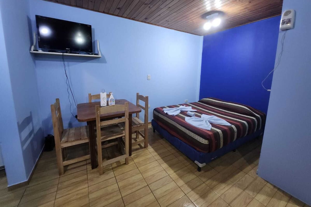 Una habitación con techo de madera, televisor montado en la pared, cama con manta a rayas, mesa de madera con sillas y pared azul.