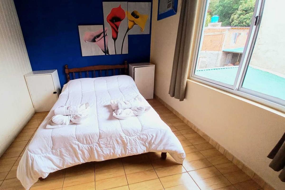 Una cama bien hecha con toallas, una pared azul con una obra de arte, una ventana con vistas y un mueble blanco pequeño.