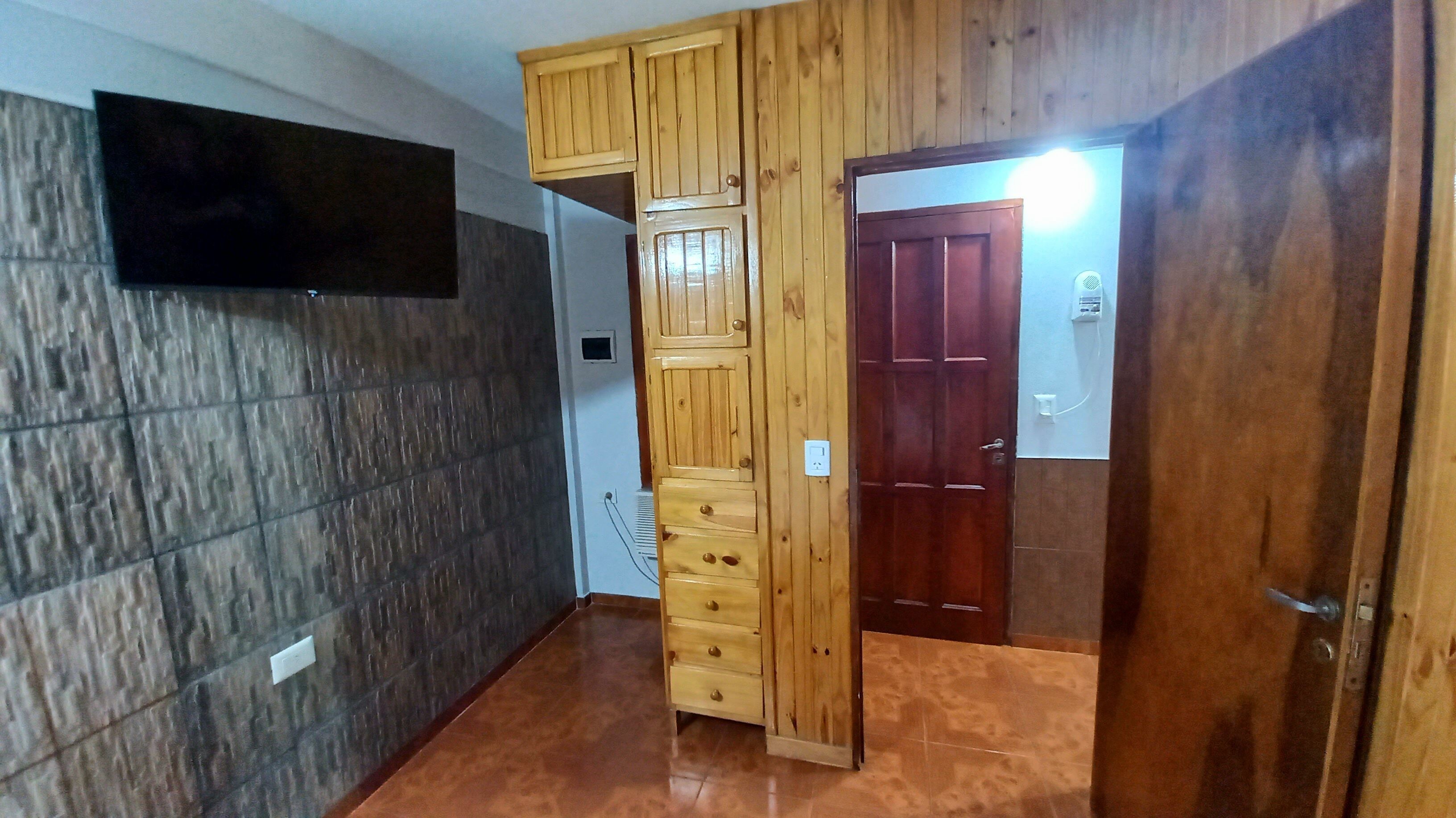 Un couloir avec une armoire en bois, un téléviseur fixé au mur et une porte surmontée d’une lampe.