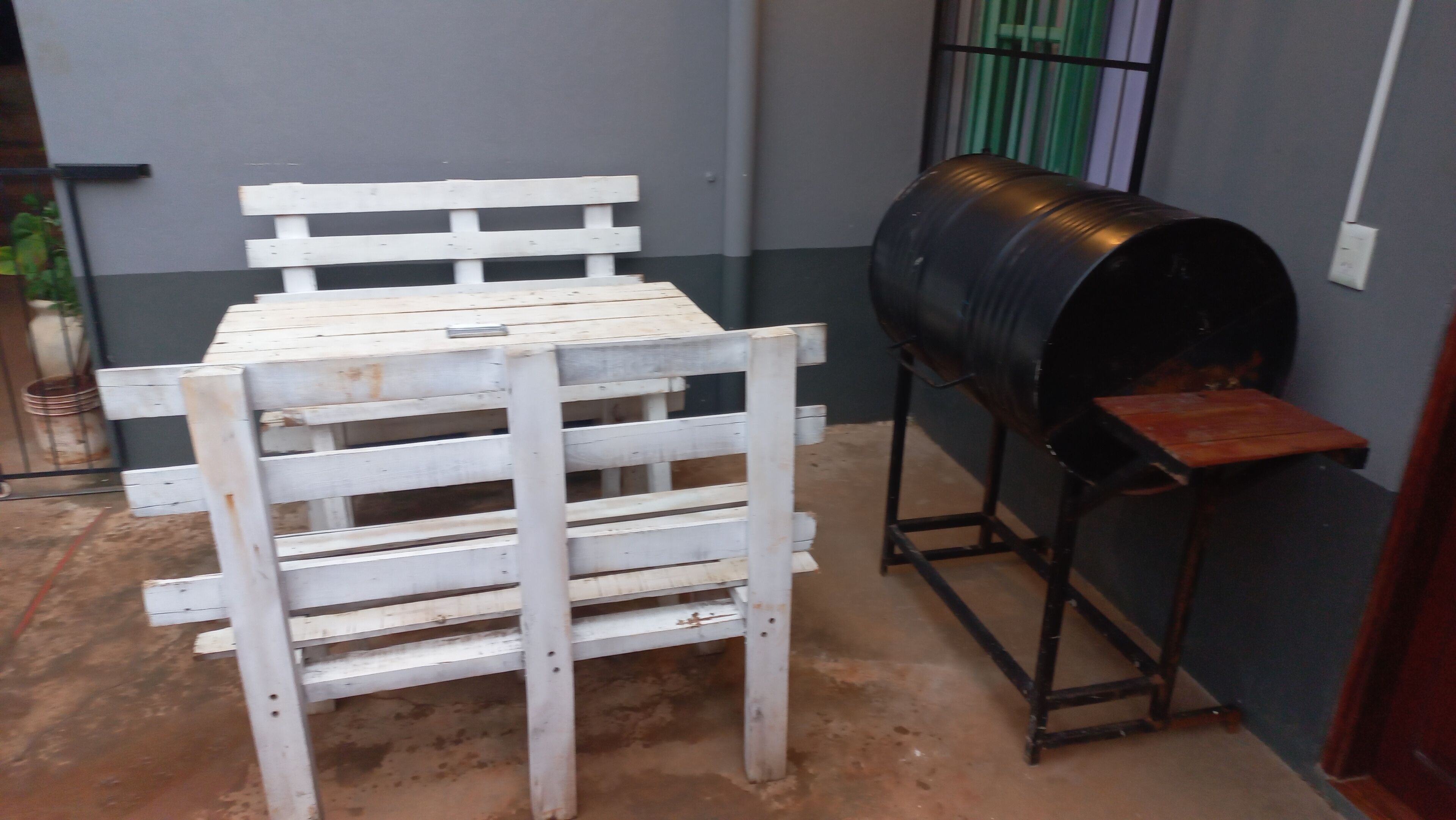 Un banco de madera blanca con una rejilla metálica fijada a una pared, un barril negro y una pequeña mesa de madera.