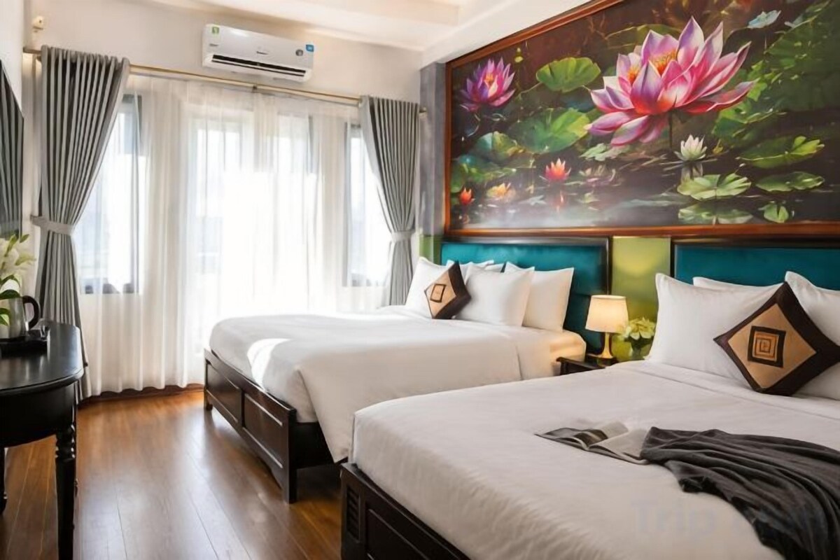 A hotel room with two beds, a painting of lotus flowers, a nightstand with a lamp, and a window with curtains.