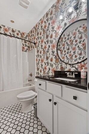 A bathroom with a white toilet, a bathtub, a sink with a black countertop, and a mirror. The walls have a floral wallpaper pattern. - DerbyGrove: Newly Renovated & Near Derby (Jeffersonville)
