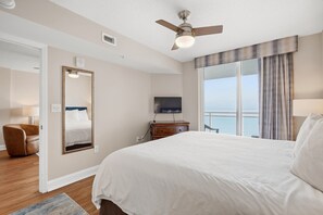 Una camera d'albergo con un letto, una televisione, una ventola a soffitto, un'ampia finestra con tende e vista sul mare.