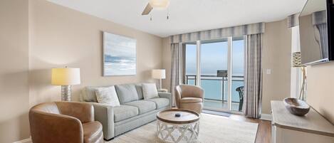 A modern living room with a ceiling fan, a sofa, two armchairs, a coffee table, and a view of the ocean through large windows.