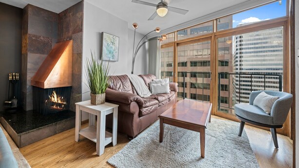 A modern living room with a fireplace, a ceiling fan, a large window with a city view, and a cozy seating area. - Condo in the Sky - Amazing Views - Gym (denver)