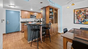 A modern kitchen with wooden cabinets, a central island with stools, a dining table, and a large abstract painting. - Condo in the Sky - Amazing Views - Gym (denver)