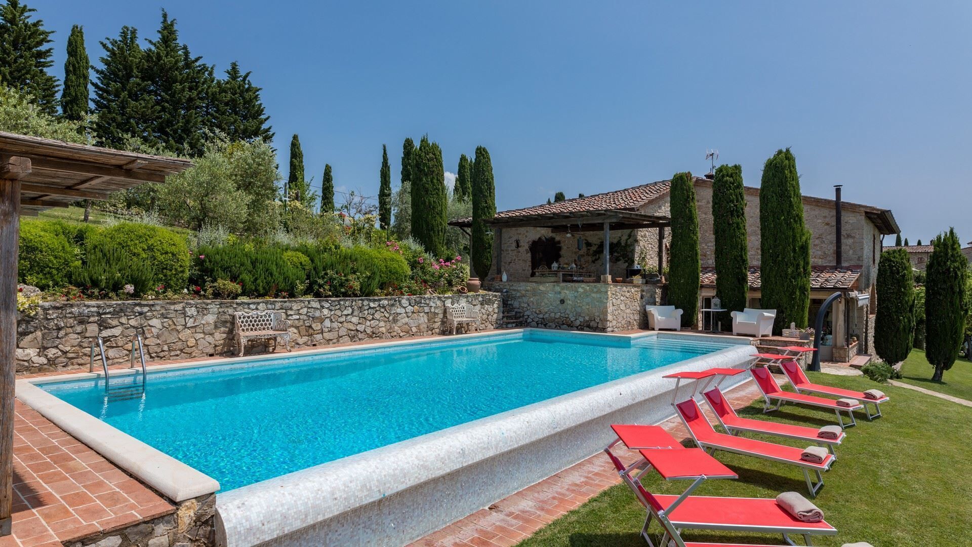 Una piscina con un muro in pietra e un pergolato su un lato, e un’area relax con sedie rosse dall’altro.