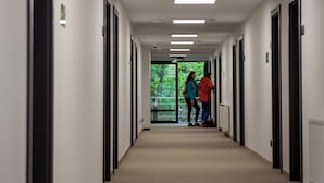 A long, narrow hallway with multiple doors on both sides, leading to rooms. There are fluorescent lights on the ceiling, and a door at the end of the hallway opens to a balcony with a view of trees. - Sport Rooms Spała (Inowlodz)