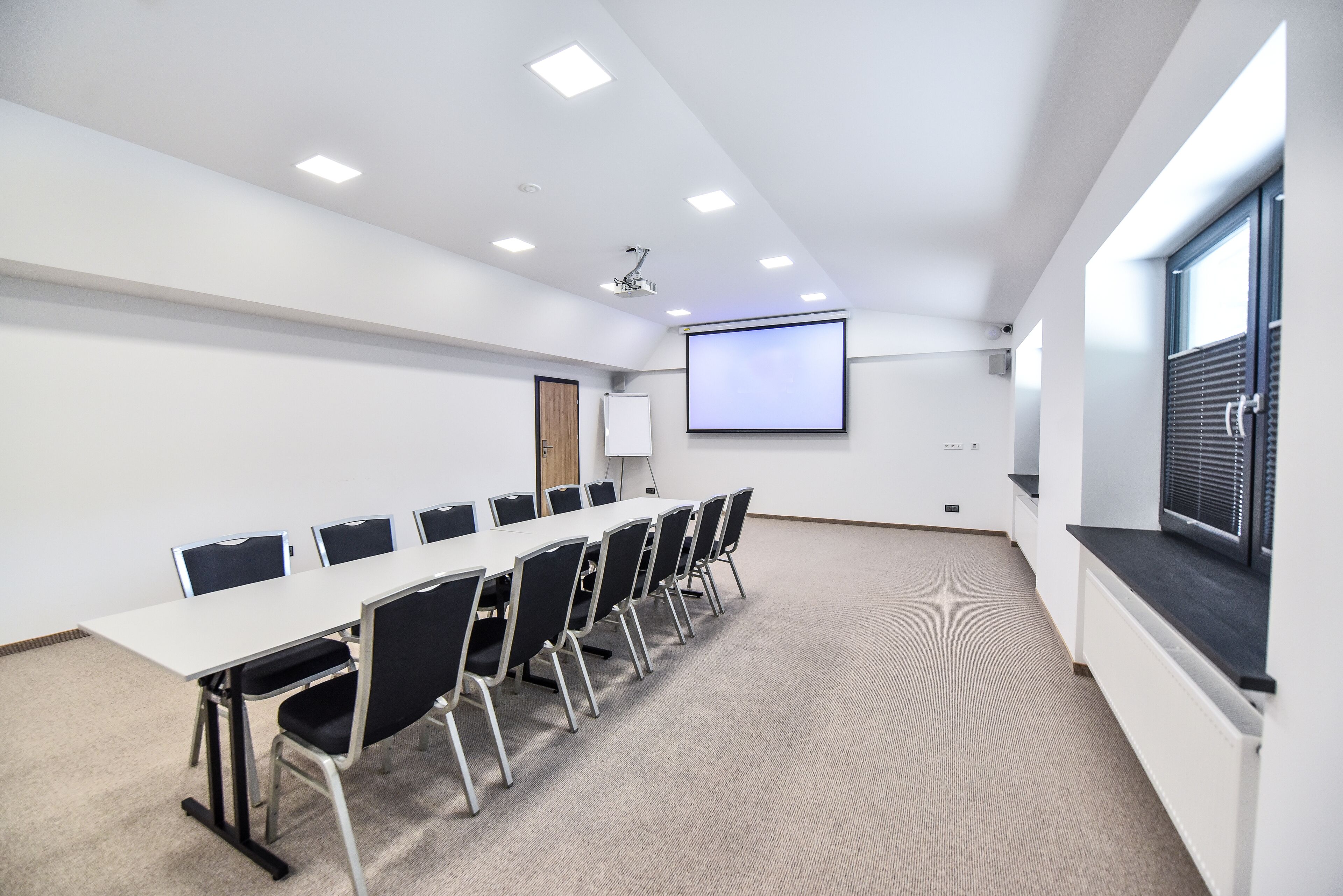A conference room with a long table, black chairs, a projector, a screen, and a window with blinds.