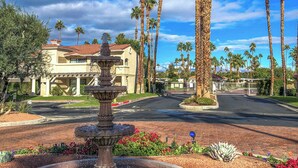 A fountain with multiple tiers, surrounded by a landscaped garden with red flowers and pebbles. Tall palm trees and a building with a red-tiled roof are visible in the background. - Cozy Ranch-Style Condo - A Ryson Property (Palm Springs)