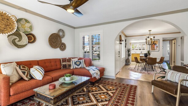 A spacious living room with a red sofa, a coffee table, and a patterned rug. There is a ceiling fan, a dining area with chairs, and a kitchen in the background. - Cozy Ranch-Style Condo - A Ryson Property (Palm Springs)