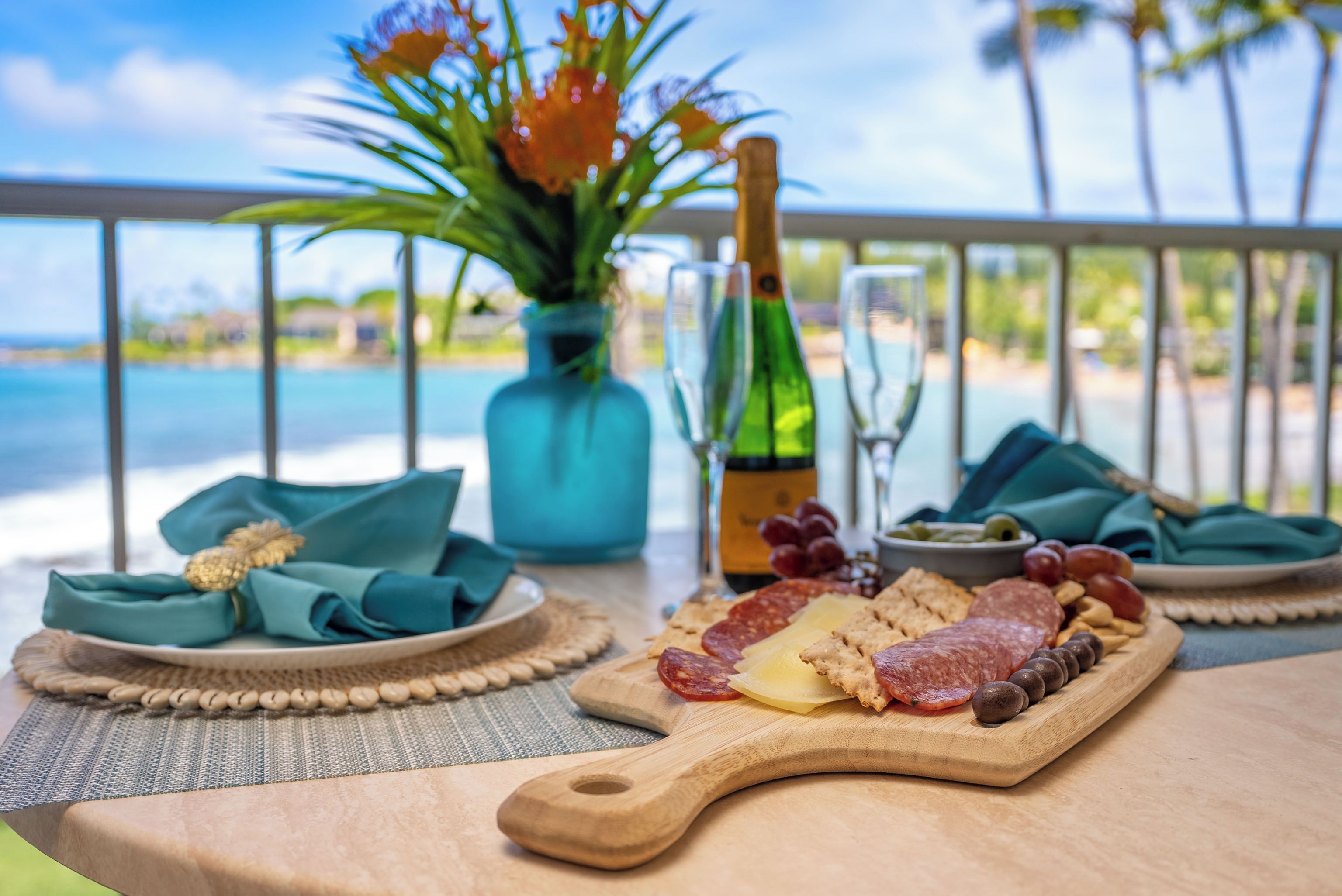 Une table avec une planche de charcuterie, une bouteille de champagne et un vase de fleurs, le tout placé sur un balcon donnant sur une étendue d’eau.