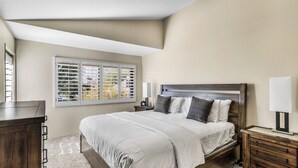 A bedroom with a large bed, a nightstand, a dresser, and a window with blinds. - Classic Comfort in Central PS - A Ryson Property (Palm Springs)