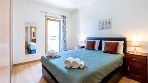 Una cama bien hecha con una colcha verde azulada y almohadas marrones, una mesita de noche de madera con una lámpara, una ventana con cortina y un espejo en la pared.